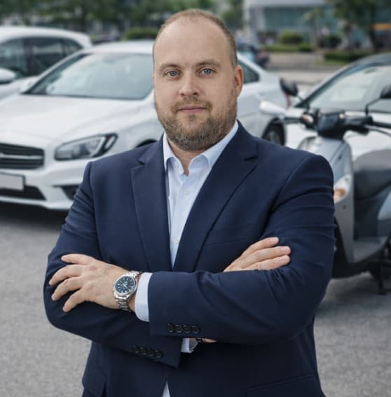 Homem barbudo de terno azul e braços cruzados posando em um estacionamento com carros.